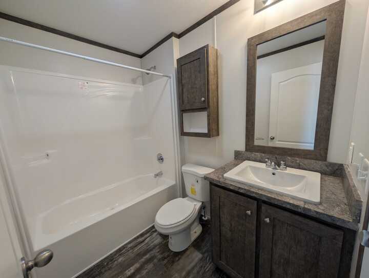 Modern bathroom with a white bathtub and shower combo, a toilet, a sink with a dark wood vanity, a matching wall cabinet, and a large mirror above the sink. The floor has a wood-look finish.