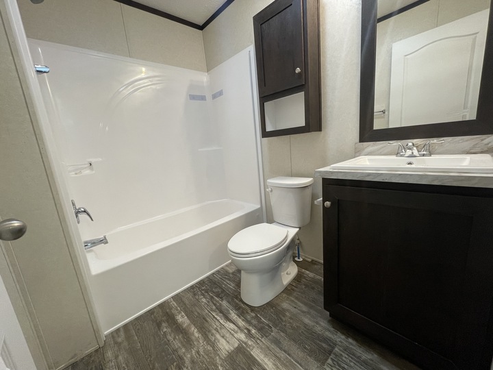 A modern bathroom featuring a white bathtub with shower, a toilet, a sink with a dark wood cabinet, a matching wall cabinet, and wood-look flooring. The walls are light-colored and the room is well-lit.