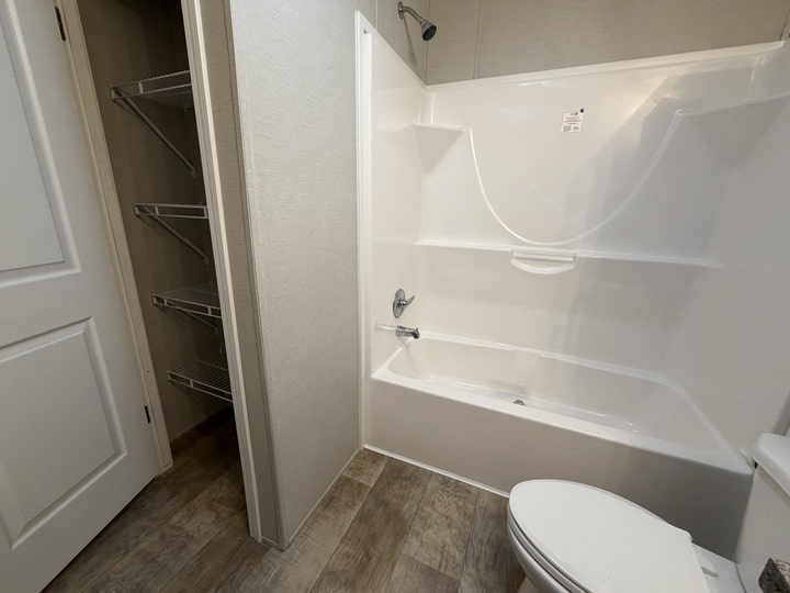 A bathroom with a white bathtub-shower combo, a white toilet, beige walls, wood-look flooring, and an open door revealing wire shelving in a closet.