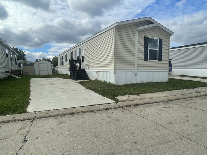A beige manufactured home with black shutters and steps sits next to a concrete driveway on a paved street. Another similar home and a small shed are visible in the background under a partly cloudy sky.