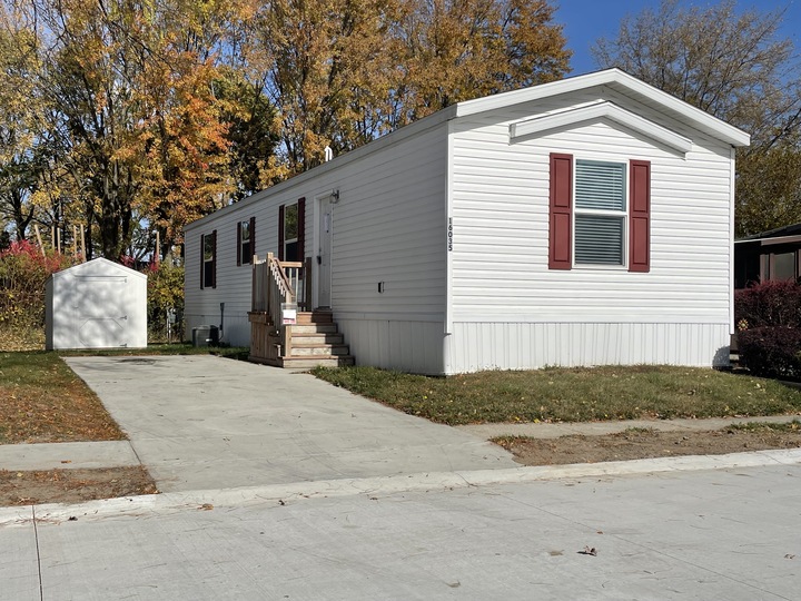 A white manufactured home with red shutters sits on a small lot with a concrete driveway and steps leading to the front door. Trees with autumn leaves and a small white shed are in the background.