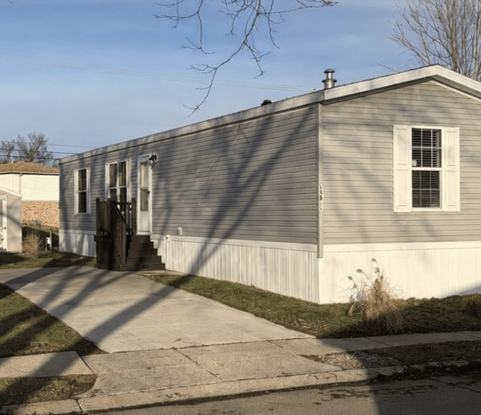 A light gray mobile home with white trim sits on a concrete driveway, with steps leading to the front door. The yard has sparse grass and a few small plants, under a clear sky.