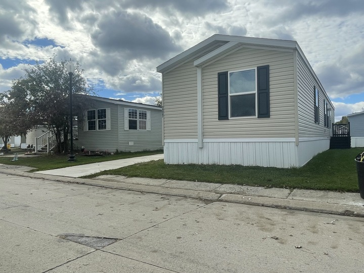 A beige manufactured home with black shutters sits on a small grassy lot beside a paved street, with another similar home and cloudy sky in the background.