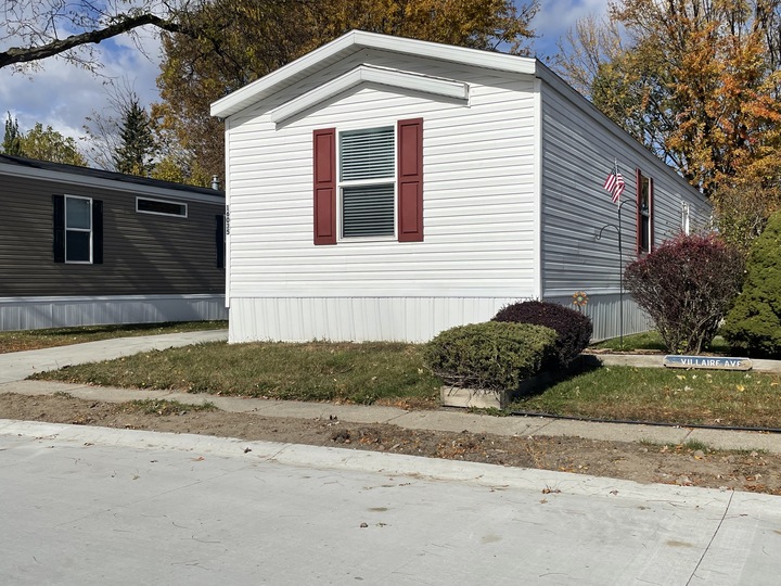 A white mobile home with red shutters, a small American flag, bushes, and a “Village Ave” street sign. The home sits beside a concrete driveway with trees showing autumn leaves in the background.