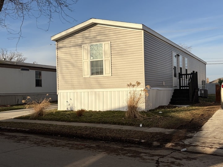 A beige manufactured home with white trim sits on a small grassy lot with ornamental grasses, next to a sidewalk under a clear blue sky. Steps lead up to a black front door on the right side of the home.