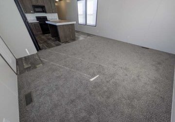 A spacious, unfurnished living room with gray carpet, adjoining a kitchen with dark wood cabinets, white countertops, a black stove, and two large windows letting in natural light.