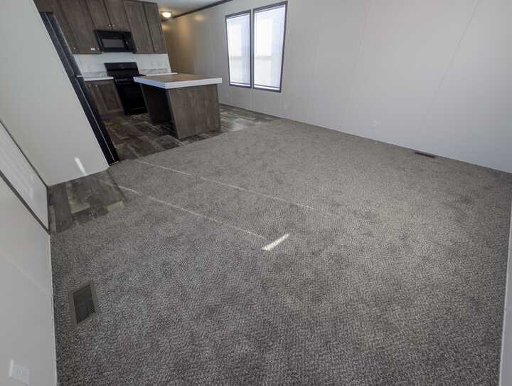A spacious, unfurnished living room with gray carpet, adjoining a kitchen with dark wood cabinets, white countertops, a black stove, and two large windows letting in natural light.