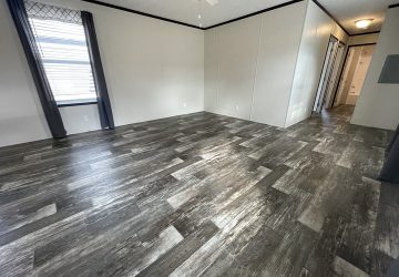 Spacious empty room with gray wood-look flooring, off-white walls, a window with gray curtains, ceiling fan with light, and a hallway leading to other rooms.