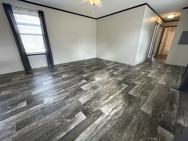 Spacious empty room with gray wood-look flooring, off-white walls, a window with gray curtains, ceiling fan with light, and a hallway leading to other rooms.