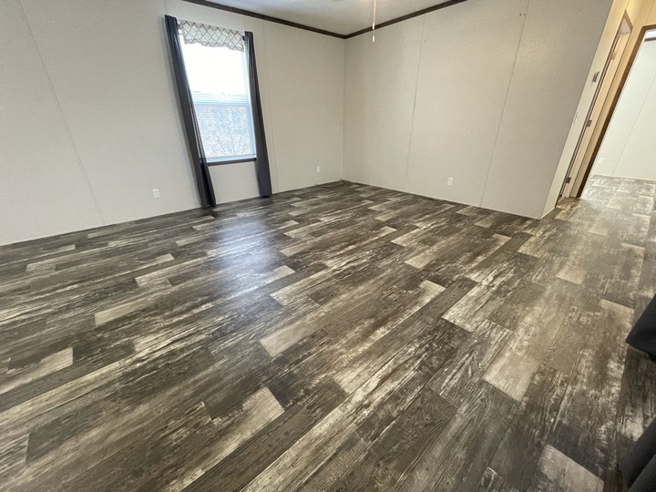 A spacious, empty room with wood-patterned vinyl flooring, beige walls, and a single window with dark curtains. Sunlight streams in, highlighting the clean, modern space. An open doorway leads to an adjoining area.