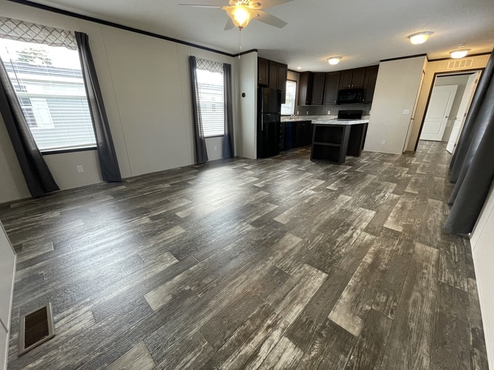 Spacious open-concept living area with wood-style flooring, large windows with dark curtains, ceiling fan, and a modern kitchen featuring dark cabinets, black appliances, and bright overhead lighting.