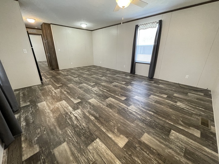 Empty room with wood-patterned vinyl flooring, cream-colored walls, a window with gray curtains, and a ceiling fan with a light. The space is well-lit with ceiling lights and natural light from the window.