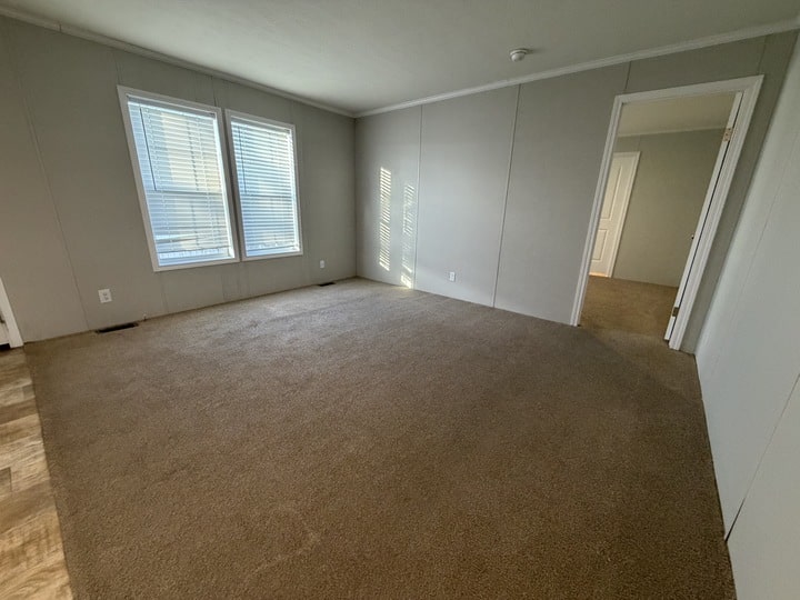 Empty room with beige carpet, light gray walls, two windows with blinds letting in sunlight, and an open doorway leading to another carpeted room. The space appears clean and well-lit.
