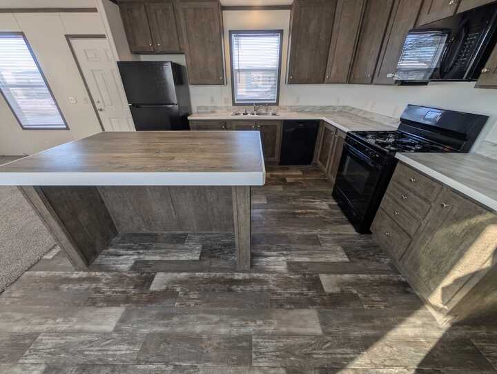 Modern kitchen with wood cabinets, a central island, black appliances including a stove, refrigerator, and microwave, and a window above the sink. The floor features wood-look tile. Bright, natural lighting fills the space.