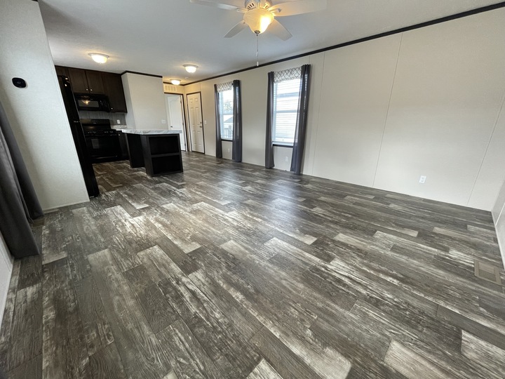 Spacious room with wood-look vinyl flooring, neutral walls, two large windows with curtains, and an open kitchen with dark cabinets and appliances in the background. A ceiling fan with a light is mounted on the ceiling.