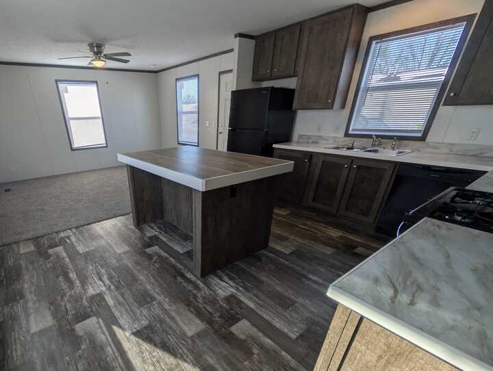 Modern kitchen with wood cabinets, a central island, dark appliances, marble-style countertops, and large windows letting in natural light. The floor has a wood-patterned finish and there is a ceiling fan in the adjacent room.