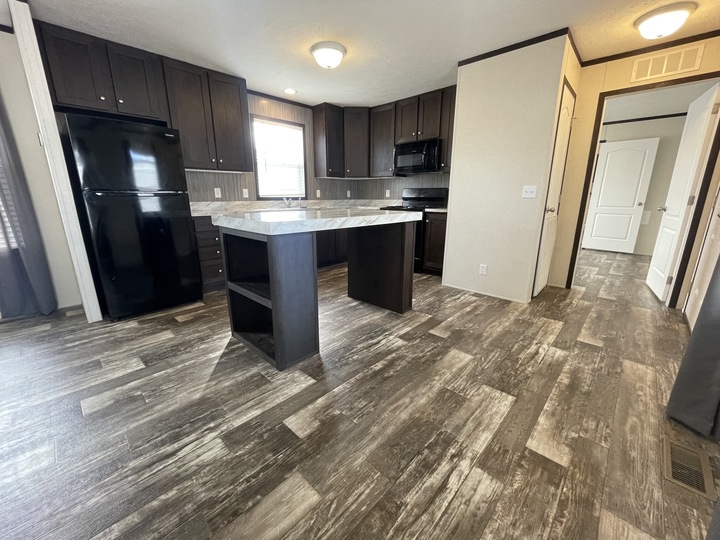 Modern kitchen with dark wood cabinets, black appliances, a central island with shelves, and gray wood-patterned flooring. A window above the sink lets in natural light. Doors to other rooms are visible.