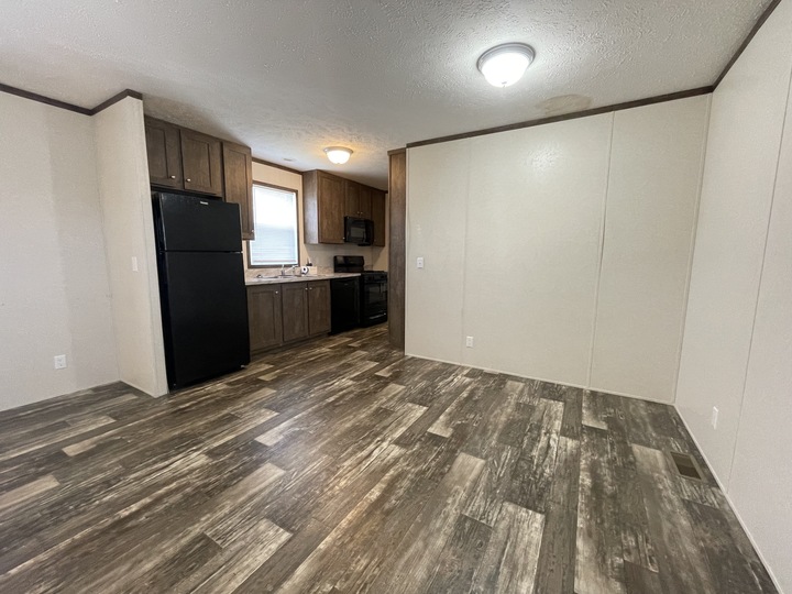 Empty room with wood-style flooring, beige walls, and a ceiling light. The kitchen area has dark cabinets, a refrigerator, a stove, a microwave, a sink, and a window above the sink.