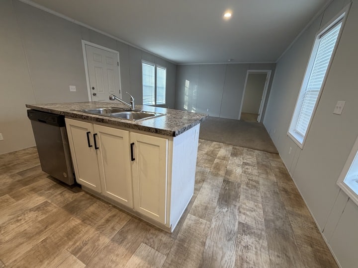 A modern kitchen with a double sink island, dishwasher, and white cabinets opens to a spacious, unfurnished living area with wood-style flooring and large windows.
