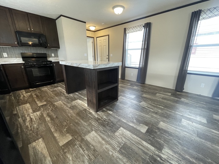 A modern kitchen with dark wood cabinets, black appliances, a marble-patterned island, and wood-look flooring, adjacent to a bright dining area with large windows and gray curtains.
