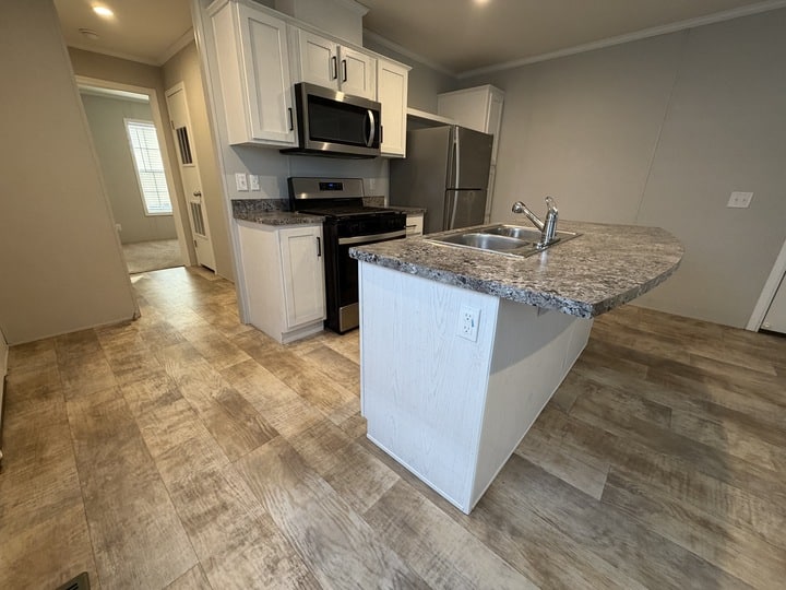 Modern kitchen with light wood cabinets, stainless steel appliances, granite-style countertops, and an island with a sink. Light wood-look vinyl flooring and neutral-colored walls create a bright, clean space.