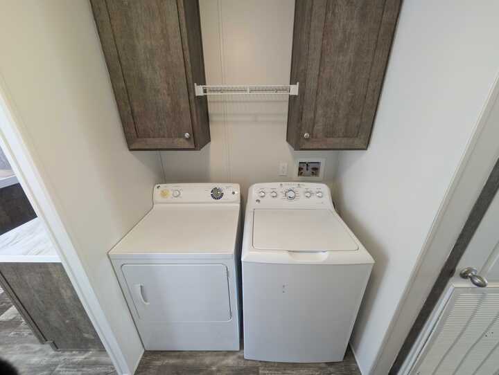 A laundry area with a white dryer and washing machine side by side, brown wooden cabinets above them, and a small wire shelf between the cabinets against a white wall.
