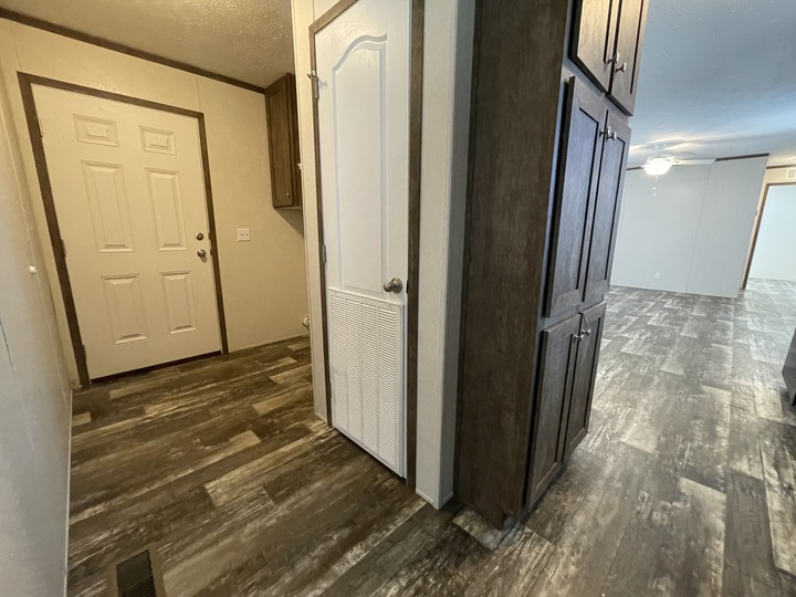 A hallway in a home with wood-patterned flooring, a beige entry door on the left, a white door with vent in the center, and dark wood cabinets on the right. The open space leads to another room in the distance.