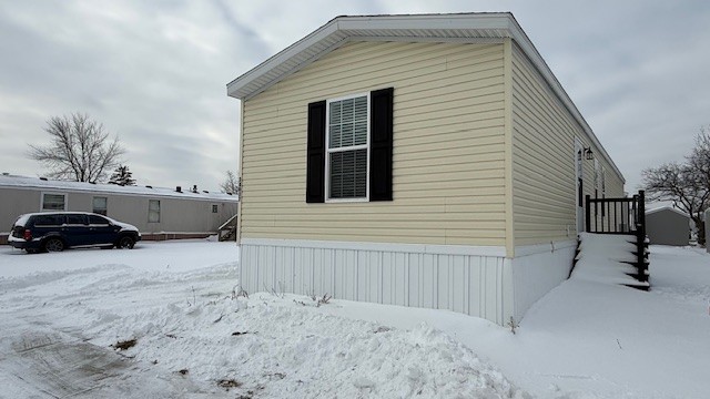 A beige manufactured home with black shutters and a white foundation sits in a snowy yard. A set of steps leads to the entrance. Snow covers the ground, and another home and a parked SUV are visible in the background.
