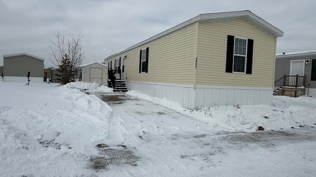 A beige mobile home with black shutters sits on a snow-covered lot. Snow is shoveled from the driveway leading to the steps. A small shed and leafless tree are visible in the background under a cloudy sky.