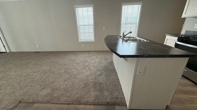 A kitchen with a curved island featuring a sink and faucet, next to an empty carpeted living area. Two windows with white blinds are visible on the back wall, and part of a stove and countertop are on the right.