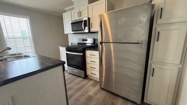 Modern kitchen with stainless steel appliances, including a refrigerator, microwave, and gas stove. Light-colored cabinets, wood-style flooring, and a double sink on a dark countertop are visible. Natural light enters through a window.