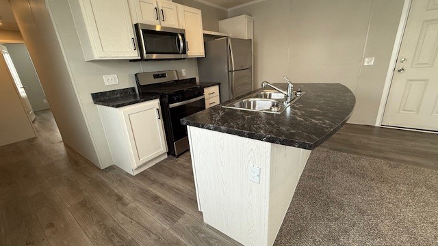 Modern kitchen with light wood cabinets, black marble countertops, stainless steel appliances, dual sink, and wood flooring. The kitchen island faces a door and carpeted living area. Hallway visible to the left.