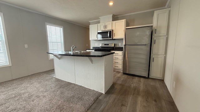 Modern kitchen with white cabinets, stainless steel appliances, an island with a sink, and a mix of carpet and wood flooring. Large windows let in natural light.