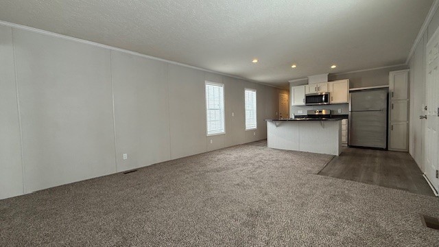 Open living area with beige carpet, two windows, and a modern kitchen featuring white cabinets, stainless steel appliances, and wood-look flooring. Ceiling lights brighten the neutral-toned space.