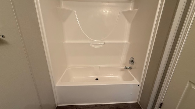 White bathtub and shower combination with a built-in soap dish and curved wall accents, set against light-colored walls and dark wood flooring. A silver faucet and drain are visible.
