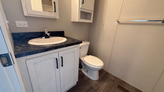 A small bathroom with a white vanity and sink, black countertop, white toilet, wall-mounted cabinet, towel bar, and wood-style flooring. The walls are light-colored and there is a vent on the floor.