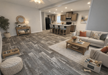 Open-concept living room and kitchen with wood-look flooring, neutral-toned furniture, a beige sofa, armchair, coffee table, and pet accessories including a toy basket and food bowls; kitchen has wooden cabinets and a breakfast bar.