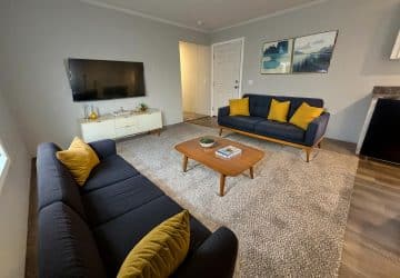 A modern living room with two navy sofas featuring yellow pillows, a wooden coffee table, a TV mounted on the wall, a white media console, gray carpet, and framed artwork above one sofa.