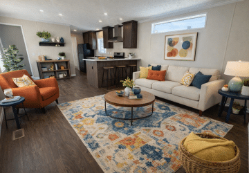 A modern living room with a white sofa, orange armchair, colorful rug, round wooden coffee table, and open kitchen in the background. Decorative pillows, plants, bookshelves, and bright artwork add cozy touches.