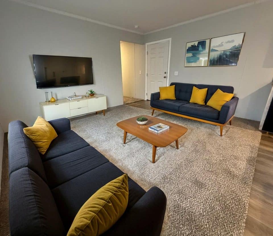 A modern living room with two navy sofas featuring yellow pillows, a wooden coffee table, a TV mounted on the wall, a white media console, gray carpet, and framed artwork above one sofa.