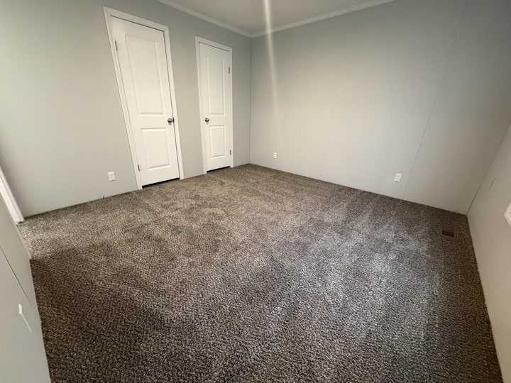 An empty room with light gray walls, two closed white doors, and plush gray carpet. The room is well-lit and has a simple, clean appearance.