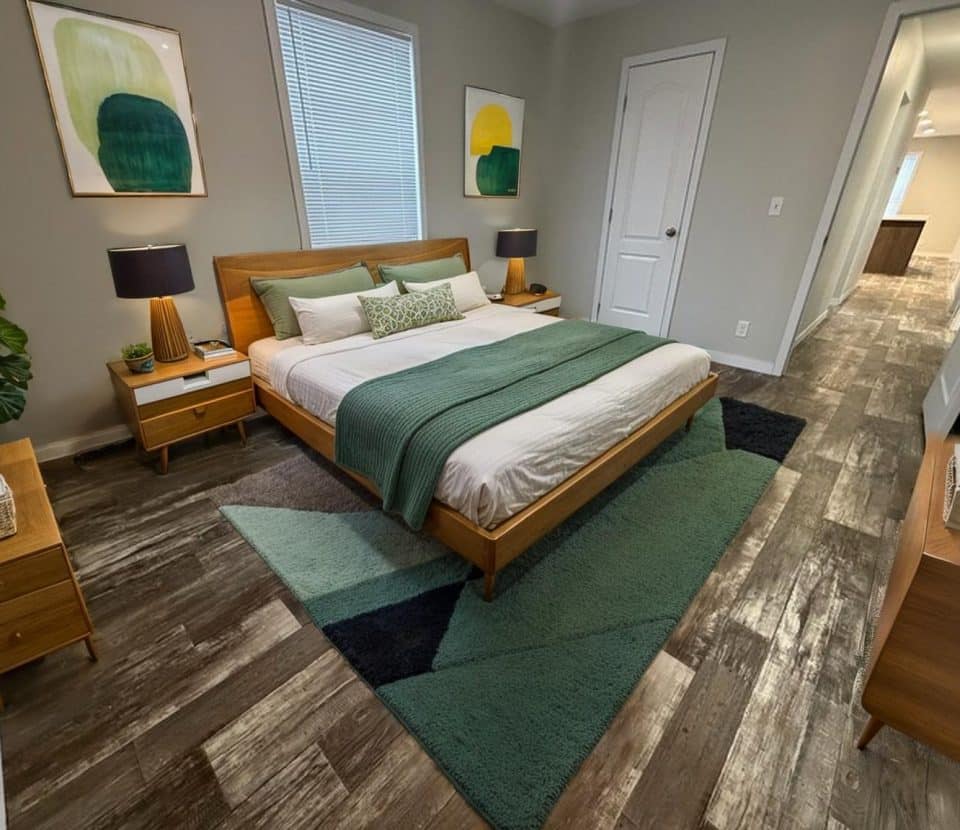 A modern bedroom with a wooden bed frame, green and white bedding, two nightstands with lamps, abstract art on the walls, a green and gray rug, and wood-look flooring. A hallway leads to a well-lit living area.