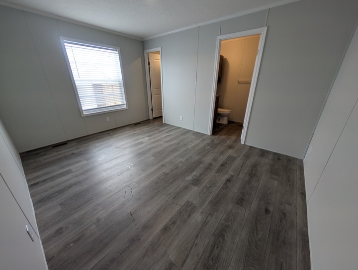 A spacious, unfurnished room with gray wood flooring, light gray walls, a large window with blinds, and two doorways—one leading to a bathroom with a visible toilet and the other to a closet.