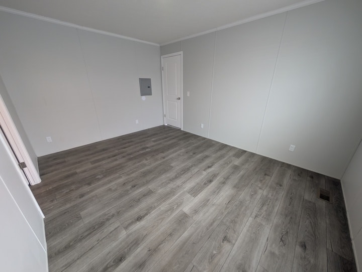 Empty room with light gray walls, white trim, and wood-style vinyl flooring. A white door and electrical panel are visible on one wall. The room is well-lit and unfurnished.