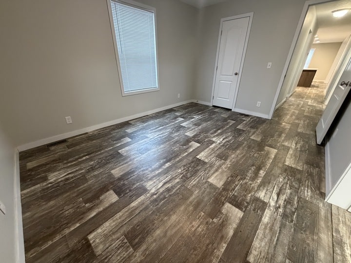 Empty room with wood-look flooring, light gray walls, a window with closed blinds, and two white doors; hallway leads to another area with similar flooring and lighting.