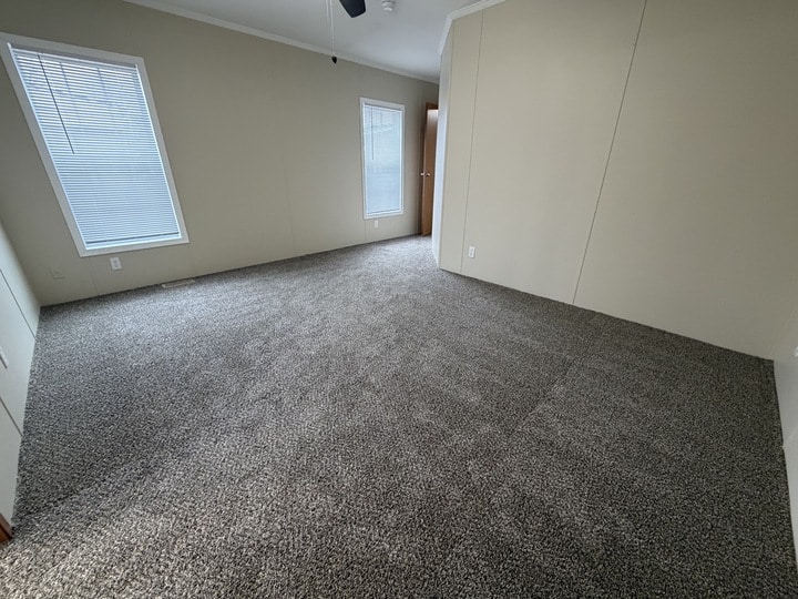 Empty room with beige walls, gray carpet, two tall windows with blinds, and a wooden door slightly open. The room appears well-lit with natural light coming through the windows.