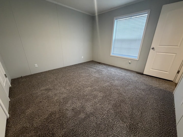 Empty room with light gray walls, brown carpet, a single window with closed blinds, and an open door. The room appears clean and well-lit, with minimal features and no furniture.