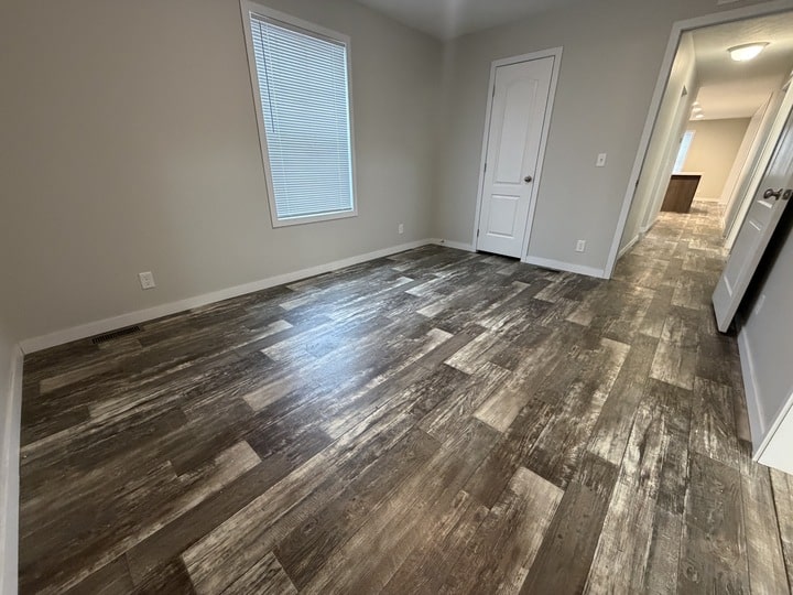 Empty room with wood-patterned vinyl flooring, light gray walls, a single window with blinds, two closed white doors, and a hallway leading to another room with ceiling lights.