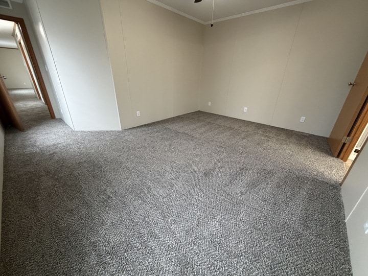 Empty room with beige walls, gray carpet, and white baseboards. An open door leads to a hallway on the left, and another doorway is visible on the right. The room appears clean and well-lit.