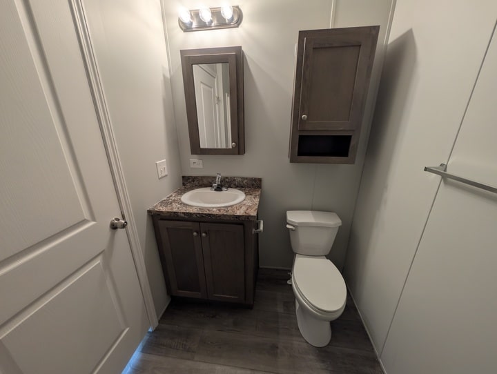 A small bathroom with a sink and cabinet, a wall-mounted mirror, a toilet, a wooden wall cabinet, towel bar, grey wood-style floor, and white walls. Three lights are above the mirror.